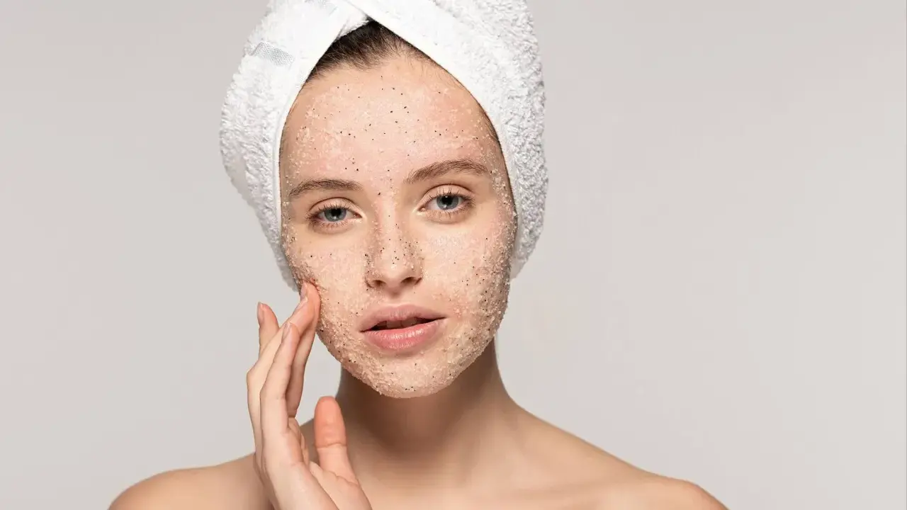 Kobieta z ręcznikiem na głowie delikatnie masuje twarz, na którą nałożyła peeling. Pokazuje, jak stosować peeling do twarzy, by uzyskać gładką skórę.