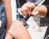 Dłoń z ciemnym lakierem do paznokci nabiera krem BB na pędzel, by nałożyć go na drugą dłoń.