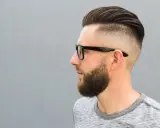 Mężczyzna w okularach z modną fryzurą na zakola, z wygolonymi bokami i zaczesaną górą. Broda dodaje mu charakteru.