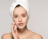 Kobieta z ręcznikiem na głowie delikatnie masuje twarz, na którą nałożyła peeling. Pokazuje, jak stosować peeling do twarzy, by uzyskać gładką skórę.