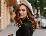 Rudowłosa kobieta w beretce i czarnym golfie, z burzą falowanych włosów i czerwoną szminką, trzyma kubek kawy.