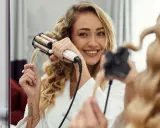 Uśmiechnięta blondynka tworzy połamane fale na włosach za pomocą lokówki.