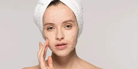 Kobieta z ręcznikiem na głowie delikatnie masuje twarz, na którą nałożyła peeling. Pokazuje, jak stosować peeling do twarzy, by uzyskać gładką skórę.