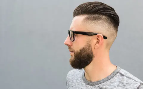 Mężczyzna w okularach z modną fryzurą na zakola, z wygolonymi bokami i zaczesaną górą. Broda dodaje mu charakteru.