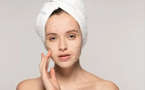Kobieta z ręcznikiem na głowie delikatnie masuje twarz, na którą nałożyła peeling. Pokazuje, jak stosować peeling do twarzy, by uzyskać gładką skórę.