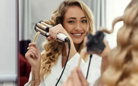 Uśmiechnięta blondynka tworzy połamane fale na włosach za pomocą lokówki.