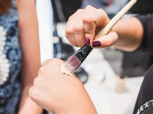 Dłoń z ciemnym lakierem do paznokci nabiera krem BB na pędzel, by nałożyć go na drugą dłoń.