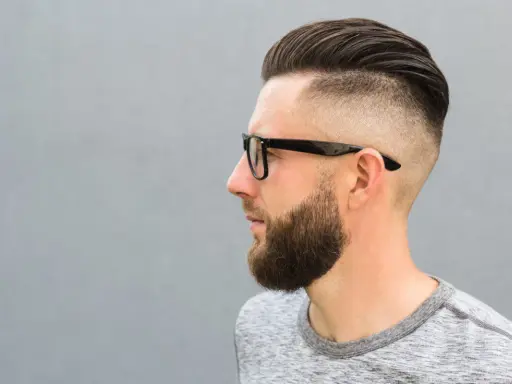 Mężczyzna w okularach z modną fryzurą na zakola, z wygolonymi bokami i zaczesaną górą. Broda dodaje mu charakteru.