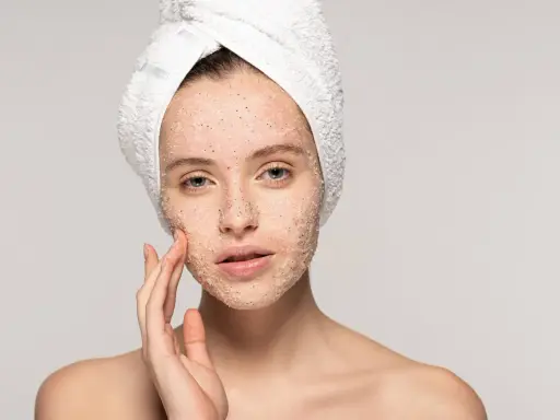 Kobieta z ręcznikiem na głowie delikatnie masuje twarz, na którą nałożyła peeling. Pokazuje, jak stosować peeling do twarzy, by uzyskać gładką skórę.