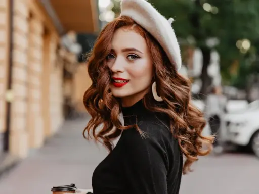 Rudowłosa kobieta w beretce i czarnym golfie, z burzą falowanych włosów i czerwoną szminką, trzyma kubek kawy.