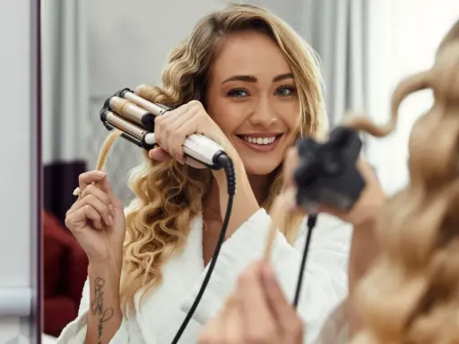 Uśmiechnięta blondynka tworzy połamane fale na włosach za pomocą lokówki.