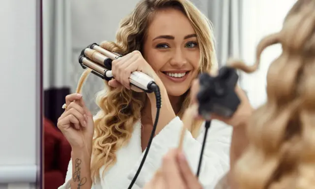 Uśmiechnięta blondynka tworzy połamane fale na włosach za pomocą lokówki.
