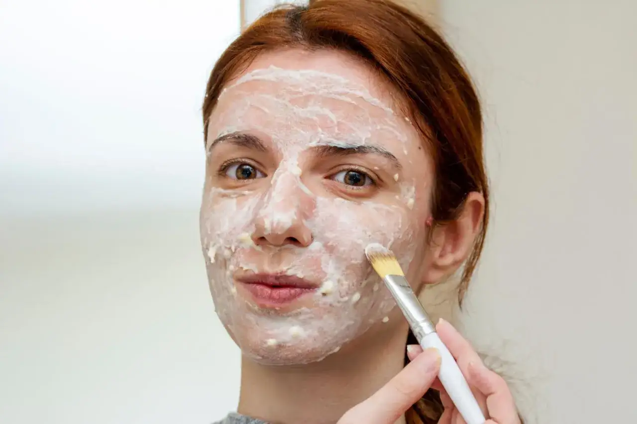 Kobieta nakłada pędzelkiem białą maskę na twarz. Dowiedz się, jak stosować peeling kwasowy dla promiennej cery.