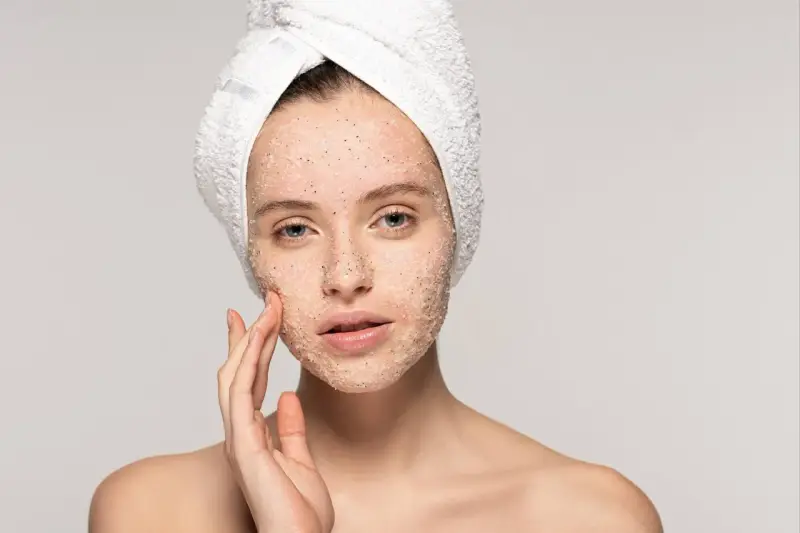 Kobieta z ręcznikiem na głowie delikatnie masuje twarz, na którą nałożyła peeling. Pokazuje, jak stosować peeling do twarzy, by uzyskać gładką skórę.